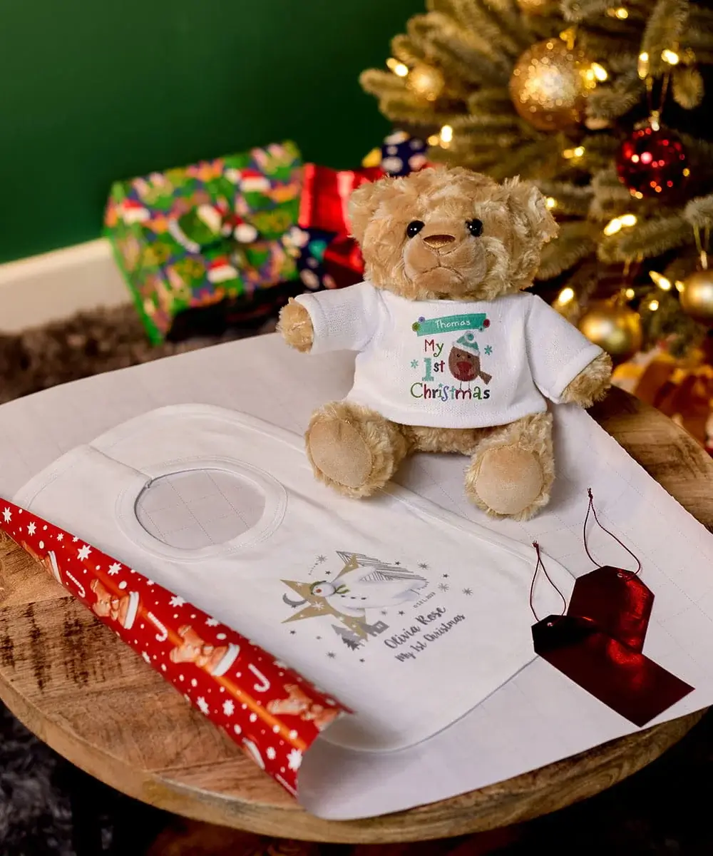 Personalised Christmas keepsake set featuring a teddy bear in a festive T-shirt, a baby handprint kit, gift wrapping paper, and Christmas decorations by a Christmas tree. Personalised Christmas keepsake set featuring a teddy bear in a festive T-shirt, a baby handprint kit, gift wrapping paper, and Christmas decorations by a Christmas tree.